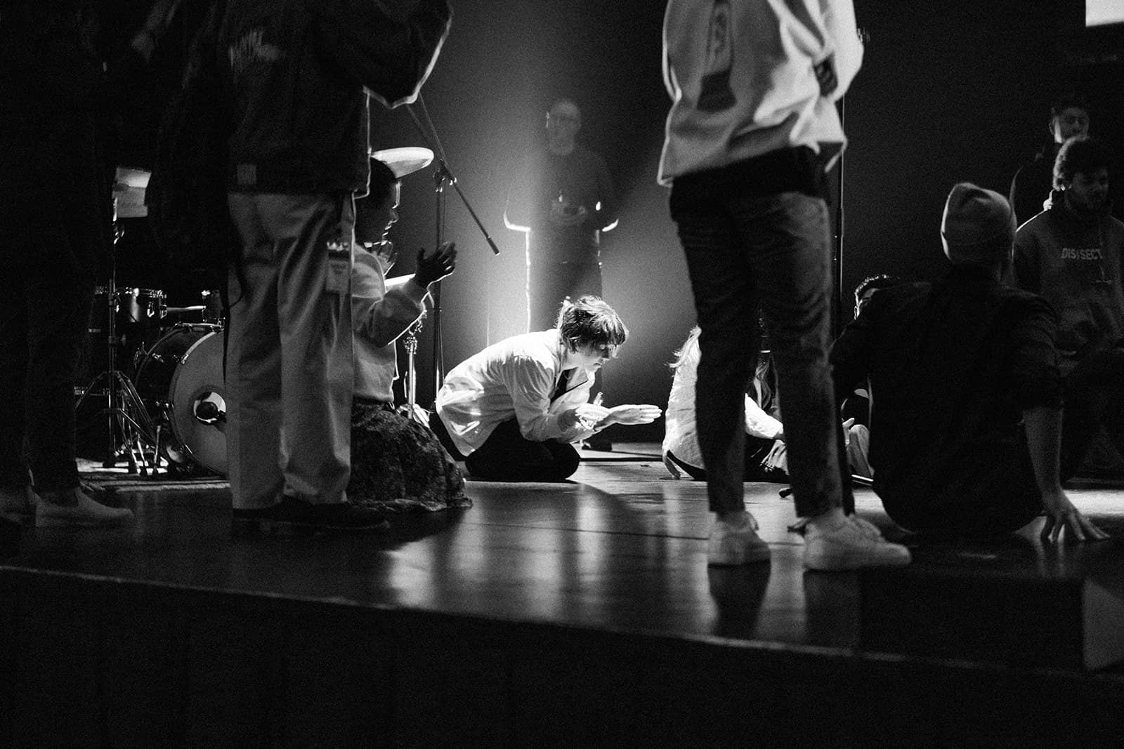 A group of people are praying near a drumset on a stage, some standing and some kneeling.