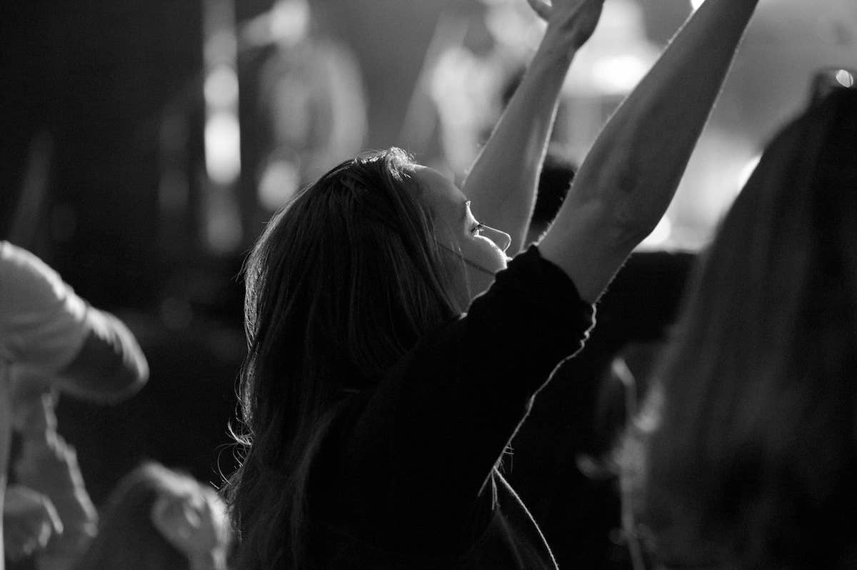 A woman with eyes closed and arms raised in worship, with other out-of-focus people around her.